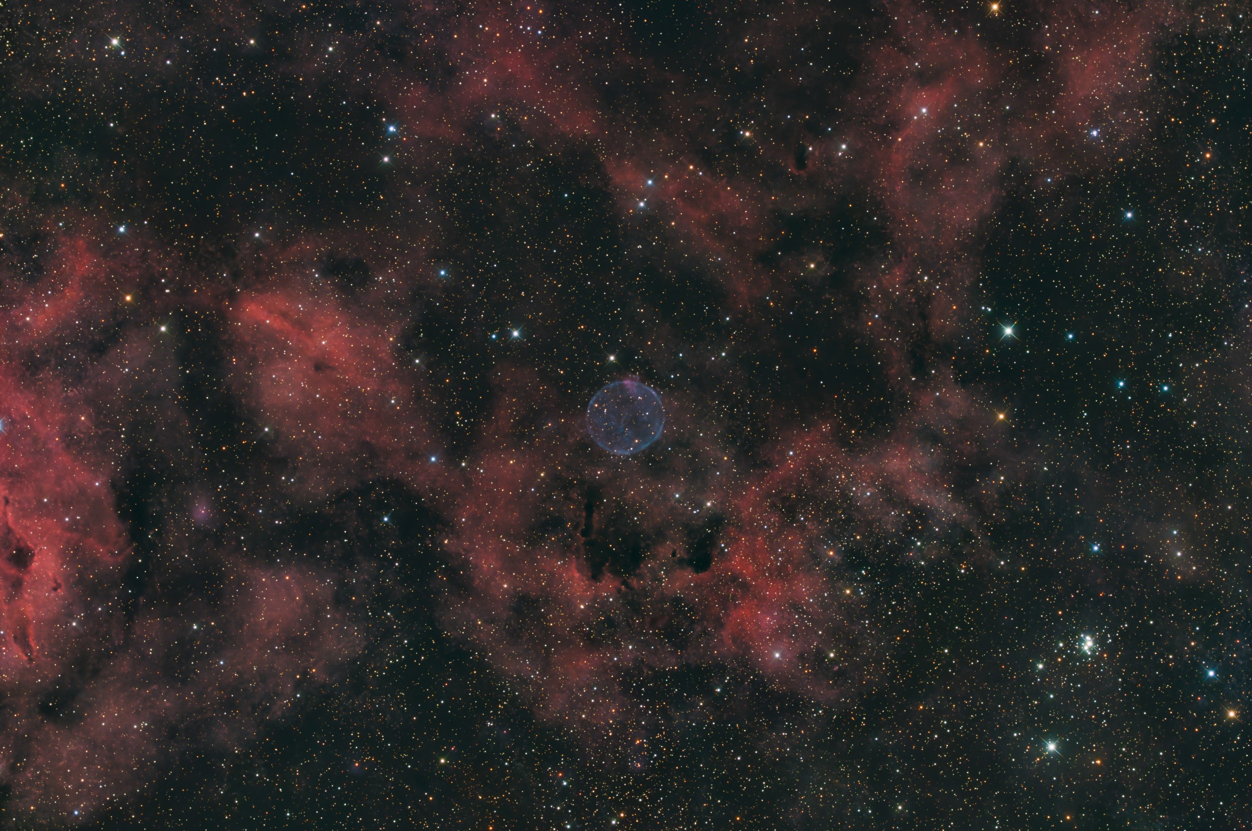 A pale blue-violet sphere glows softly near the center of a dense starfield, set against faint rust-red clouds of nebulosity. Dark, ropy dust lanes thread through the reddish haze, and a small cluster of bright blue-white stars sits in the lower right corner.