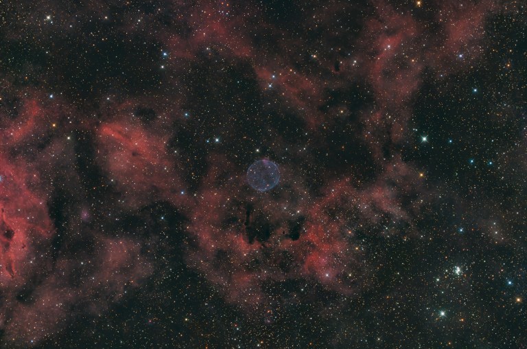 A pale blue-violet sphere glows softly near the center of a dense starfield, set against faint rust-red clouds of nebulosity. Dark, ropy dust lanes thread through the reddish haze, and a small cluster of bright blue-white stars sits in the lower right corner.