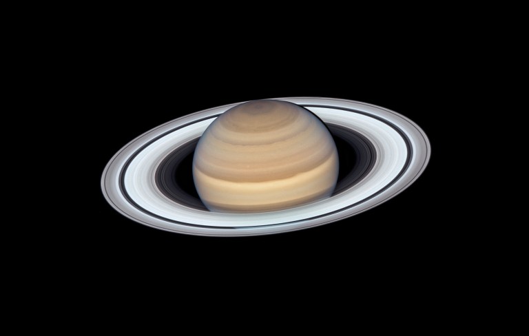 Photograph of Saturn against a black background, tilted so its northern hemisphere faces the viewer with wide ring system encircling it. The planet's disk shows subtle bands of tan, cream, and pale gold, with a darker hexagonal-tinted region visible near the north pole. The rings display distinct concentric divisions, including a prominent dark gap, and cast a soft shadow onto the upper part of the globe.