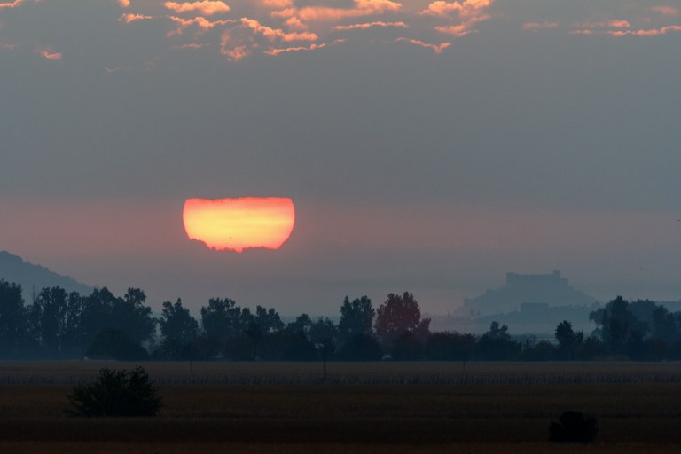 A photograph of the autumnal equinox sunrise over a hazy rural landscape in Spain, showing the Sun as a large, heavily distorted red-orange disc flattened by atmospheric refraction just above the treeline. A silhouetted tree line, distant mountains, and what appears to be a castle or fortress on a hilltop are visible through the haze. Pink-tinted clouds and a gray-blue sky fill the upper portion of the frame, and a flat golden field stretches across the foreground.