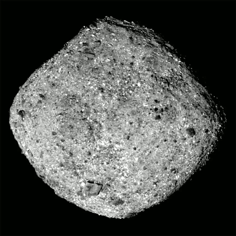 A monochrome, close-up photograph shows a diamond-shaped asteroid with a rough, gravel-like surface centered against a solid black background. Bright, direct lighting highlights thousands of tiny, reflective grey and white specks interspersed with dark, pitted shadows and scattered boulders. High contrast emphasizes its jagged, rounded perimeter and uneven, rubble-covered terrain.