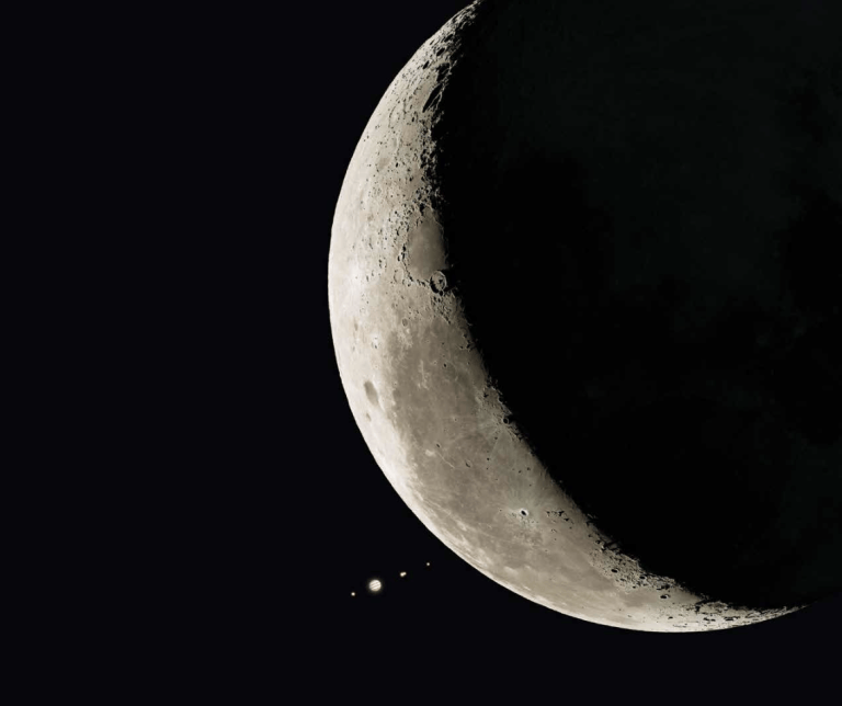 A photoillustration of a thick crescent Moon against a black sky, filling the right side of the frame. The sunlit crescent curves along the left edge, showing gray maria and many craters, and the rest of the Moon's disk is in shadow. Just off the Moon's lower-left limb sits Jupiter, a small tan disk with visible cloud bands, lined up with its four Galilean moons as tiny points of light: from left to right, Ganymede, Jupiter, Io, Callisto, and Europa. The scene shows the jovian system a few minutes before the Moon occults it. Empty black space fills the left side of the image.