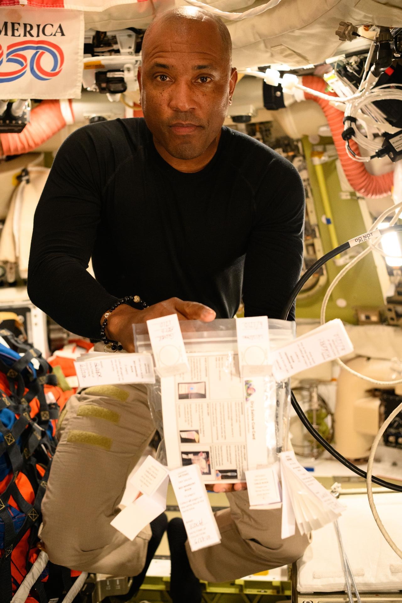 Photograph of a bald astronaut in a black long-sleeved shirt and khaki pants floating inside a cluttered space station module, looking directly at the camera. He holds a clear zip bag with a printed instruction card, with several small white paper sample strips taped around it. A partial banner reading AMERICA 250 appears at upper left, with orange ducting, white cables, and stowed gear surrounding him.