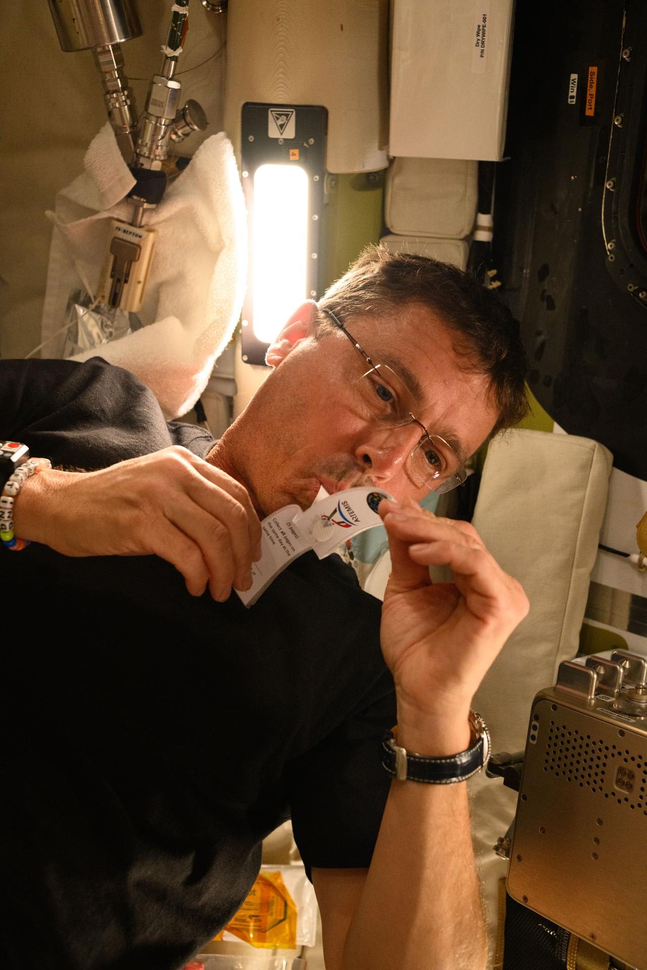 Photograph of an astronaut in glasses and a black shirt, shown sideways inside a space station module, holding a small white paper sample strip to his mouth. The strip carries an Artemis logo and printed instructions. He wears a beaded bracelet and a wristwatch, and behind him a bright light panel, white cloth-wrapped fittings, and equipment panels fill the cramped compartment.