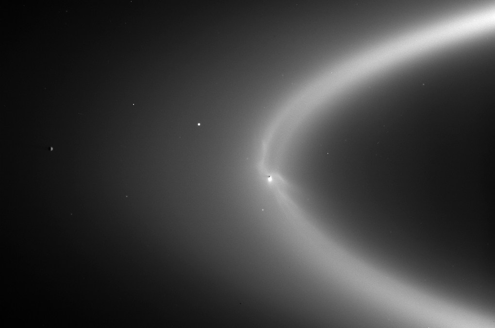Black-and-white photograph of Saturn’s faint E ring, a broad, hazy arc of light sweeping from upper right toward the center. A bright point marks Enceladus where the arc bends sharply at center, with a few small moons and stars scattered across the dark background.