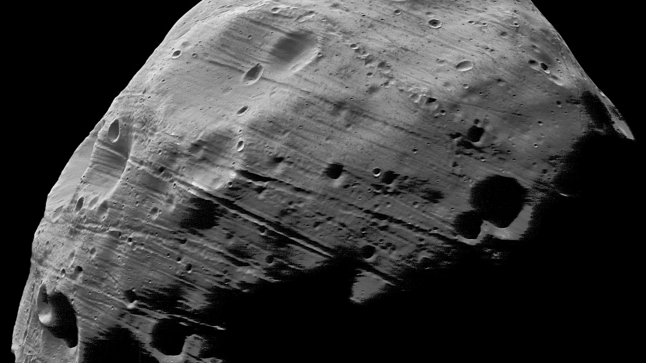 Black-and-white photograph of Mars’s moon Phobos, a lumpy, gray body filling most of the frame. Its surface is marked by small craters, long parallel grooves, and a large crater at lower left, with dark shadows across the lower right against a black background.