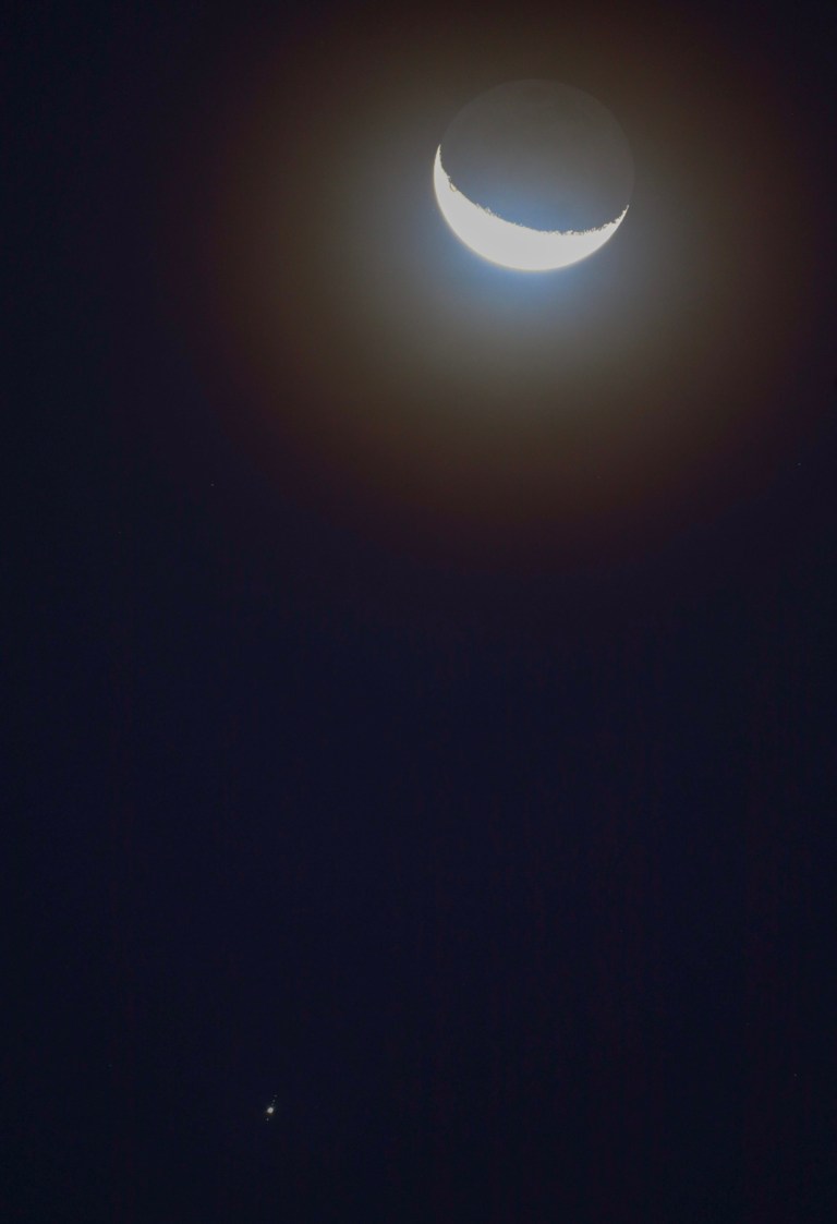 A thin, bright crescent Moon curves along the lower left edge of a pale gray disk near the top of a tall, dark navy frame, wrapped in a soft brownish glow. Far below, near the bottom left, a small bright dot sits in a short diagonal line of tiny specks, three above it and one below.