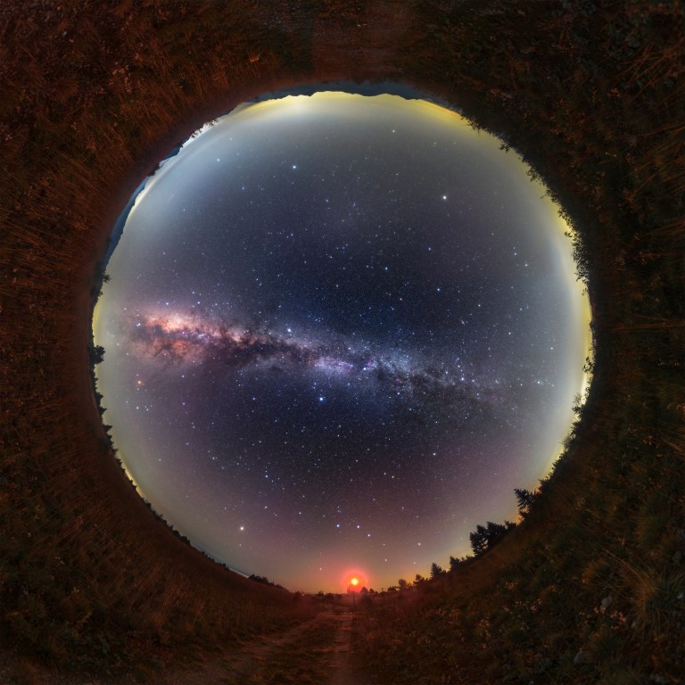 A circular all-sky photo framed by a ring of reddish grass and dark trees that curls around the edges like a tunnel. Inside, a deep blue sky full of stars is crossed from left to right by the Milky Way, a hazy band with a glowing pinkish core and dark streaks on the left. Pale yellow-gray light rims the horizon, and a small bright red light glows at the bottom above a dirt track.
