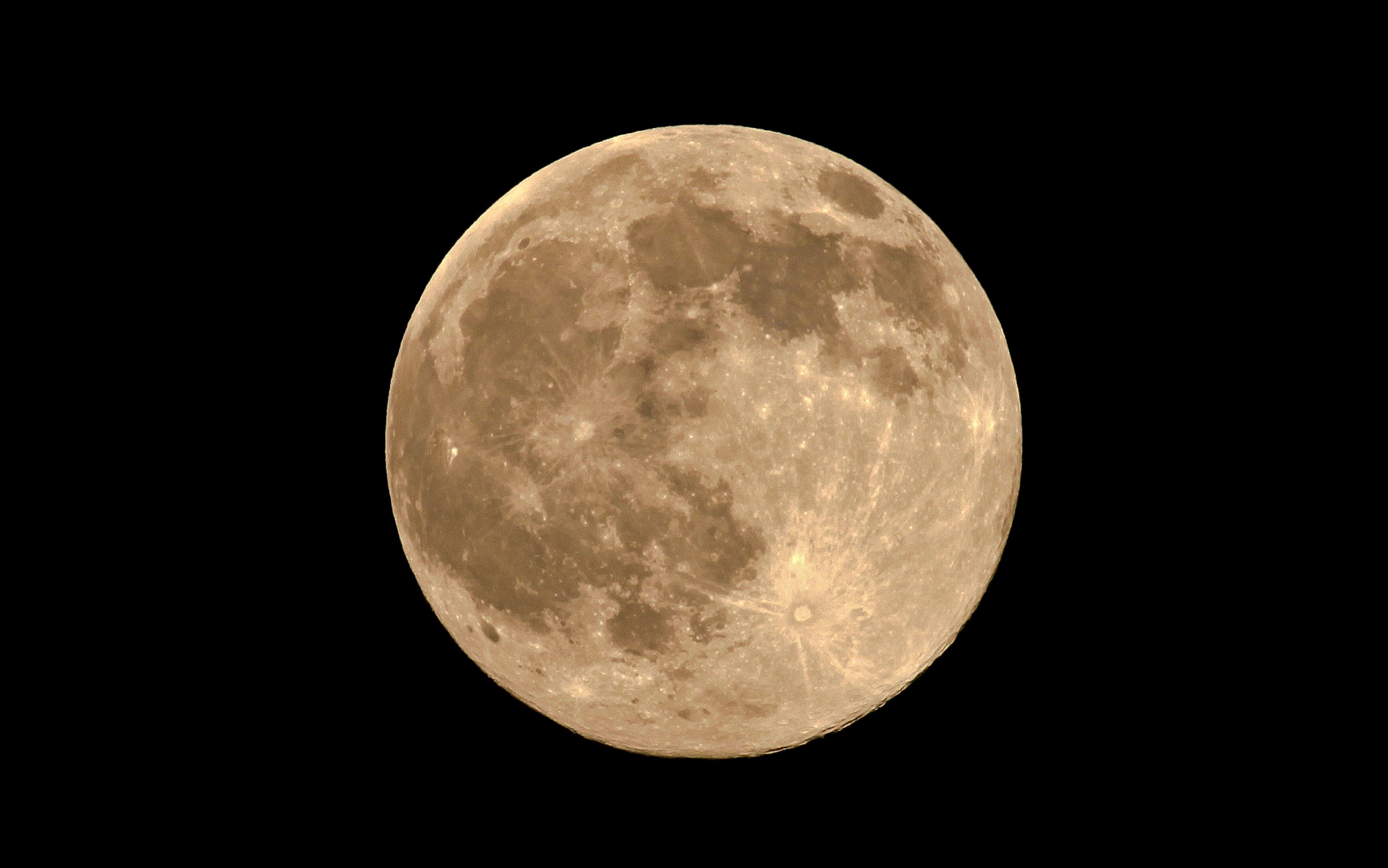Photograph of a full Moon against a black sky, tinted golden-tan. Dark gray lunar maria cover much of the upper and left areas, and bright rayed craters stand out, including a prominent one at lower right.
