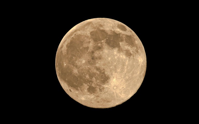 Photograph of a full Moon against a black sky, tinted golden-tan. Dark gray lunar maria cover much of the upper and left areas, and bright rayed craters stand out, including a prominent one at lower right.