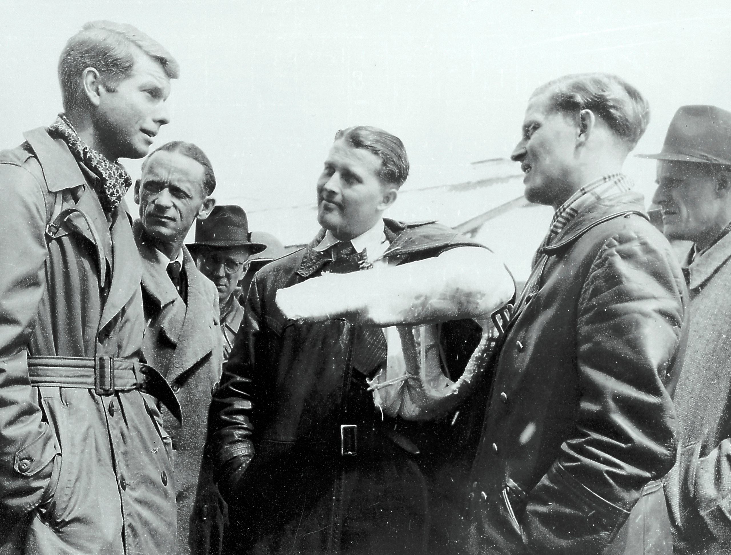 A black-and-white photograph shows a group of men standing outdoors, engaged in conversation. The central figure wears a dark leather jacket and has his left arm held horizontally in a large, white cast. To his left and right, other men in trench coats, leather jackets, and fedoras look toward him under bright, high-contrast outdoor lighting.