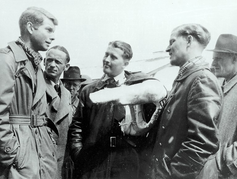 A black-and-white photograph shows a group of men standing outdoors, engaged in conversation. The central figure wears a dark leather jacket and has his left arm held horizontally in a large, white cast. To his left and right, other men in trench coats, leather jackets, and fedoras look toward him under bright, high-contrast outdoor lighting.