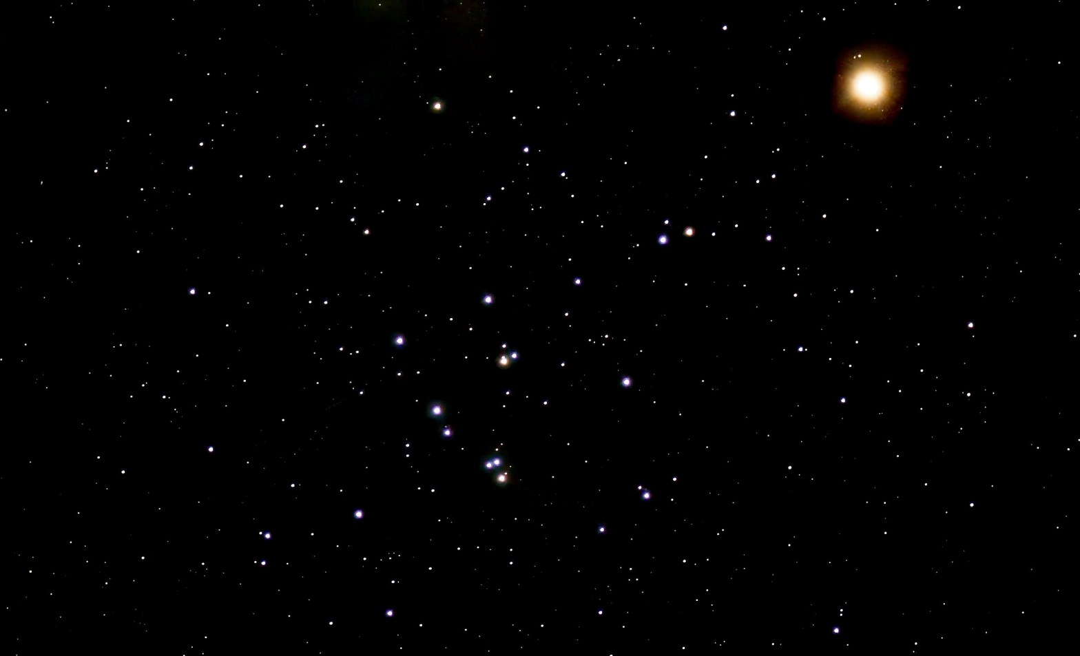 Photograph of the Beehive Cluster (M44) as a loose scattering of blue-white stars with a few orange stars across the lower center of the frame, set against a black sky filled with fainter stars. Mars appears at upper right as a bright, glowing orange-red disk.