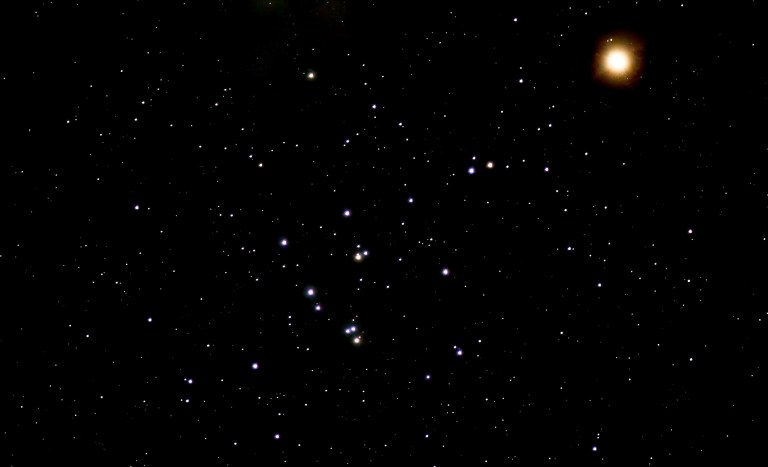 Photograph of the Beehive Cluster (M44) as a loose scattering of blue-white stars with a few orange stars across the lower center of the frame, set against a black sky filled with fainter stars. Mars appears at upper right as a bright, glowing orange-red disk.