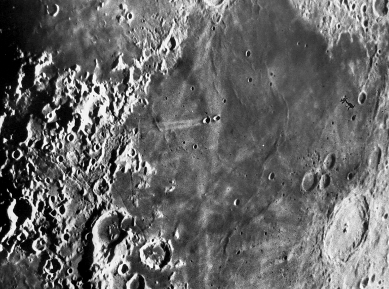 Black-and-white photograph of a region of the Moon's surface, showing a broad, dark, smooth lava plain at center bordered by rugged, heavily cratered highlands at left. Numerous small craters dot the plain, a bright ray-like streak runs near a pair of craters at center left, and a large, shallow crater with terraced walls sits at lower right. A small dark smudge or speck appears at upper right.