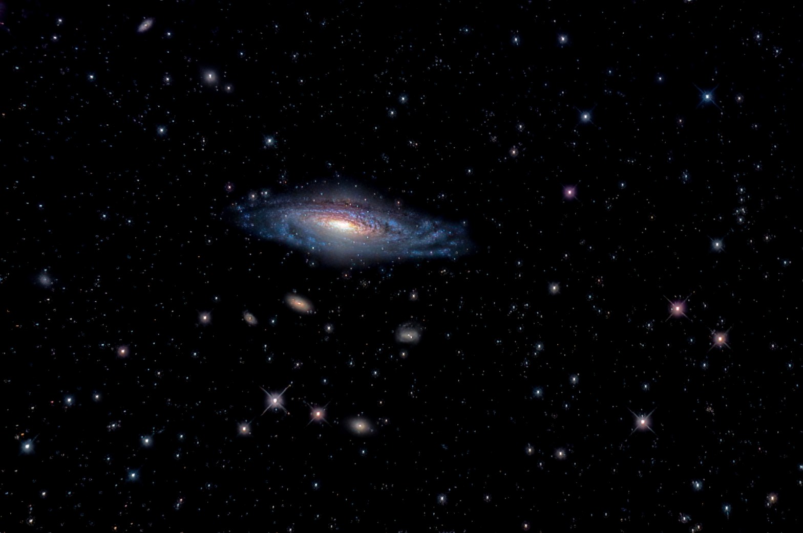 Photograph of the spiral galaxy NGC 7331, tilted diagonally at center with a bright golden core, dark dust lanes, and blue spiral arms. Several smaller, fainter galaxies lie below it, and numerous stars, some with diffraction spikes, are scattered across the black background.