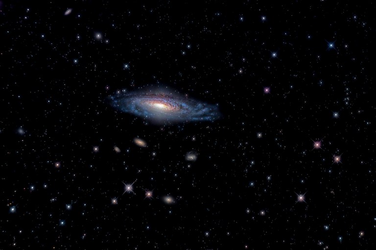 Photograph of the spiral galaxy NGC 7331, tilted diagonally at center with a bright golden core, dark dust lanes, and blue spiral arms. Several smaller, fainter galaxies lie below it, and numerous stars, some with diffraction spikes, are scattered across the black background.