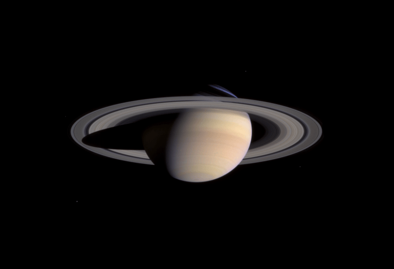 Photograph of Saturn against a black background, tilted with its northern hemisphere tipped toward the viewer and pale cream and tan cloud bands crossing its globe. The wide ring system casts a dark shadow band across the planet's upper portion, with a faint bluish tint visible at the upper edge of the rings where they cross in front of the disk.