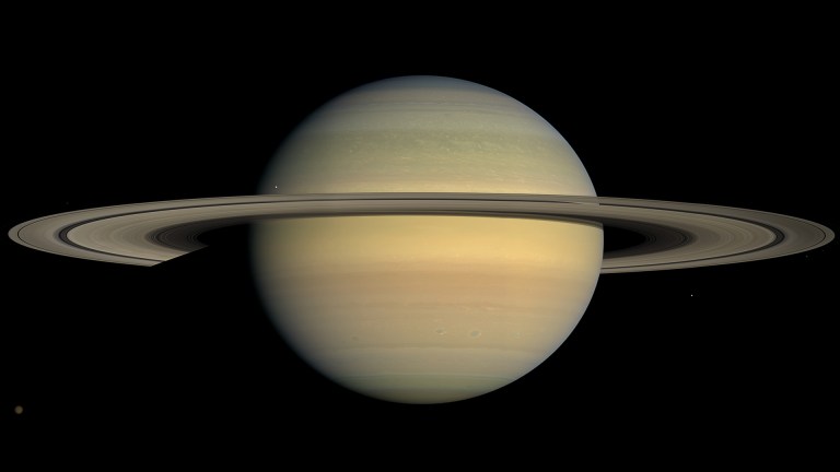 Photograph of Saturn against a black background, shown nearly face-on with pale cream and tan cloud bands crossing its globe and a faint orange tint near the equator. The wide ring system extends horizontally across the frame, and several small moons appear as faint points of light scattered around the planet.