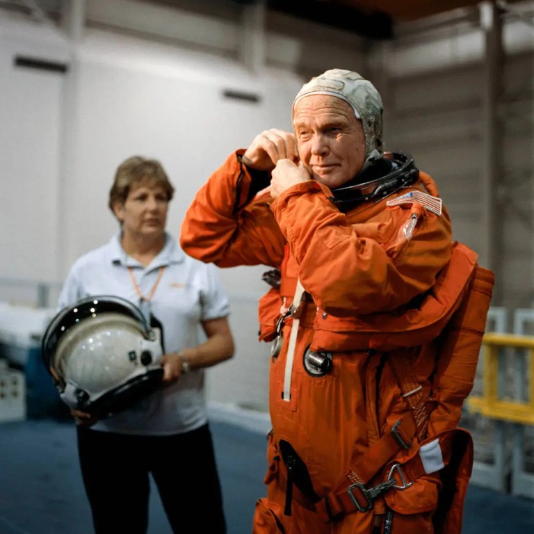 An older man wearing a bright orange flight suit and a tight white cap stands in the foreground, adjusting straps near his neck under clear indoor lighting. Behind him and slightly out of focus on the left, a woman in a light grey polo shirt stands holding a round helmet with a tinted visor. The background consists of a plain, light-colored room with neutral industrial walls and equipment.