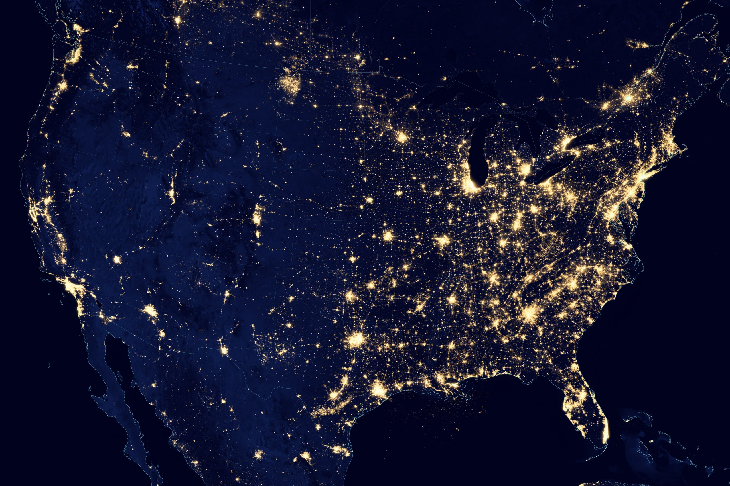 Satellite photograph of the contiguous United States at night, showing city lights as golden points and clusters against a dark navy landmass and black ocean. The eastern two-thirds of the country glow densely and almost continuously along the coasts and around the Great Lakes, while the western half shows sparser, more isolated clusters of light concentrated along the Pacific coast and scattered through the interior.