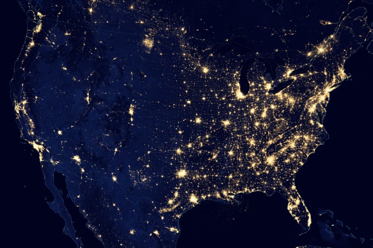 Satellite photograph of the contiguous United States at night, showing city lights as golden points and clusters against a dark navy landmass and black ocean. The eastern two-thirds of the country glow densely and almost continuously along the coasts and around the Great Lakes, while the western half shows sparser, more isolated clusters of light concentrated along the Pacific coast and scattered through the interior.