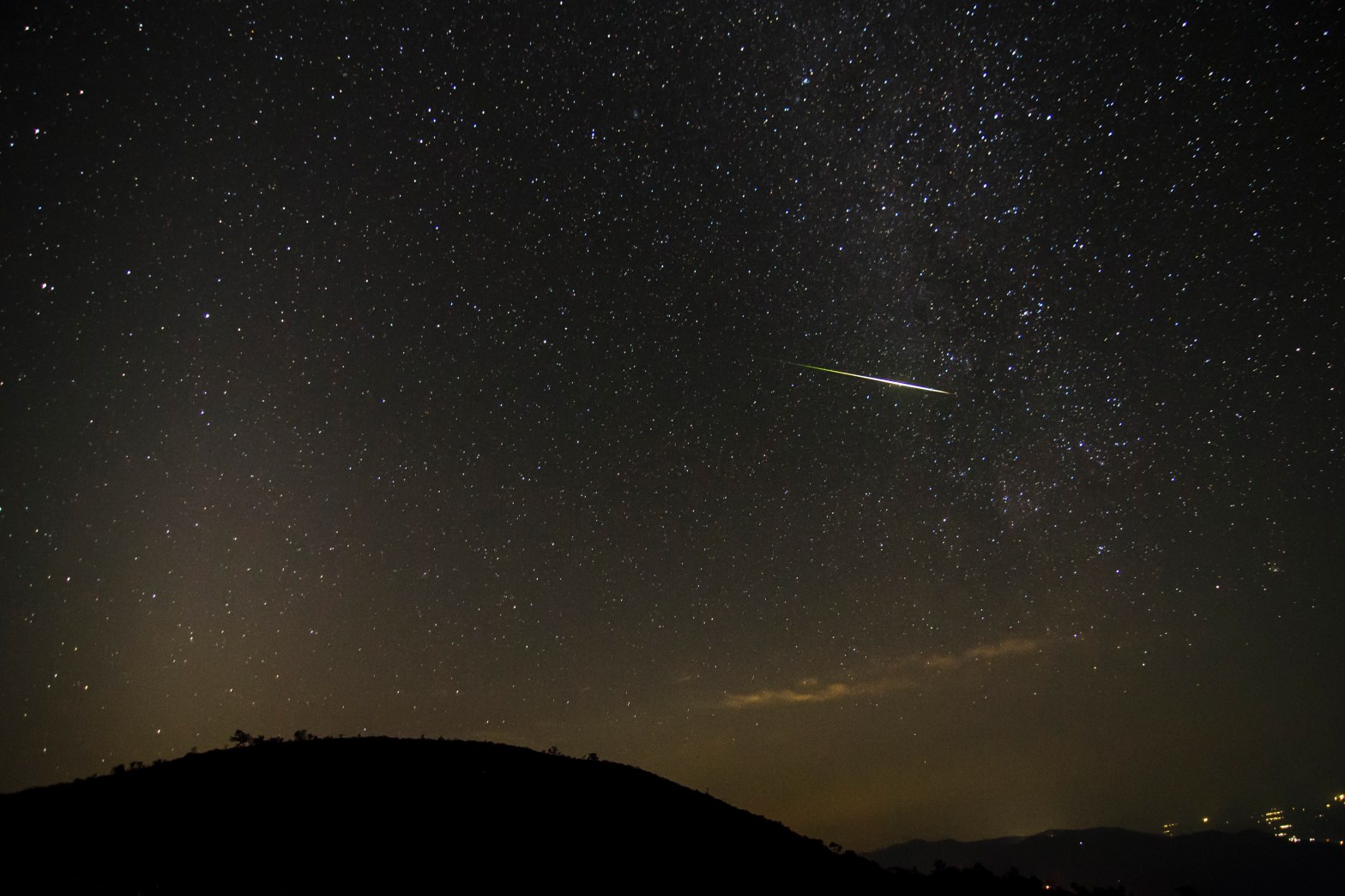 Photograph of a star-filled night sky over the dark silhouette of a hill, with a bright green-yellow Orionid meteor streaking diagonally at center right. The Milky Way is visible at upper right, a faint tan glow of zodiacal light rises from the horizon, thin clouds lie low at right, and a few distant town lights appear at lower right.
