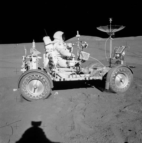 The lunar rover kicks up some dust as Dave Scott maneuvers it at the beginning of their second lunar excursion. His right hand is on the T-shaped handle used to steer the vehicle. Just in front of his hand is a traverse map that shows the Hadley Rille along its western edge and the cratered terrain to the east.