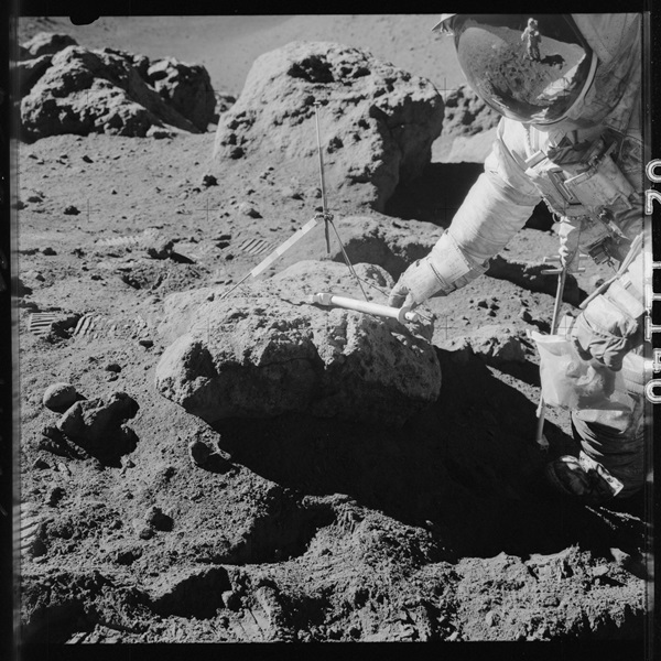 Dave Scott reaches for a hammer atop a boulder near the Hadley Rille at Station 9A. His left hand is holding a sample bag with at least one sample in it.