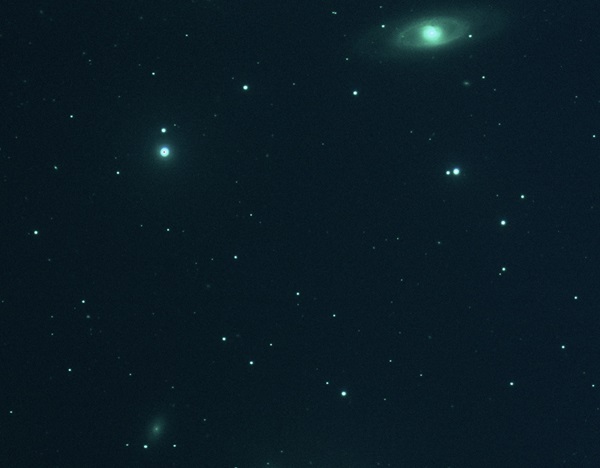 A star field with a pale green galaxy at upper right, a nebulous core surrounded by wispy spiral arms that nearly form a complete ring.