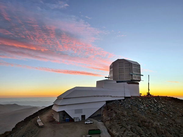 A futuristic-looking building set into the side of a mountain leads to a massive telescope dome at the summit. A post-sunset orange glow is on the horizon, illuminating cirrus clouds with pastel pinks above a landscape of misty mountains.