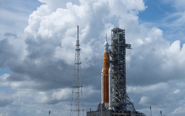 An orange rocket flanked by white booster rockets and topped with a white capsule sits on a launchpad against a sky filled with cumulus clouds
