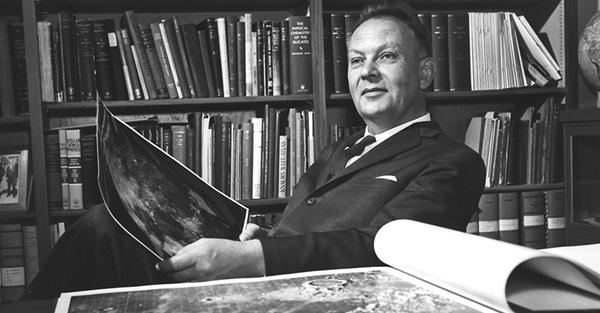 A man in a suit leans back in a chair with an atlas of the Moon in front of him and an image of the Moon in his hand. A bookshelf is behind him.