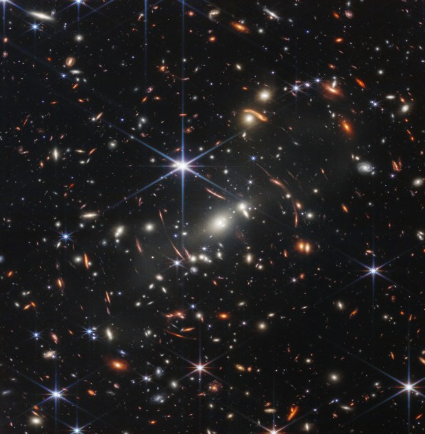 A field of bright galaxies against a black background. Some forming a central cluster and others are distorted, appearing to wrap around the cluster.