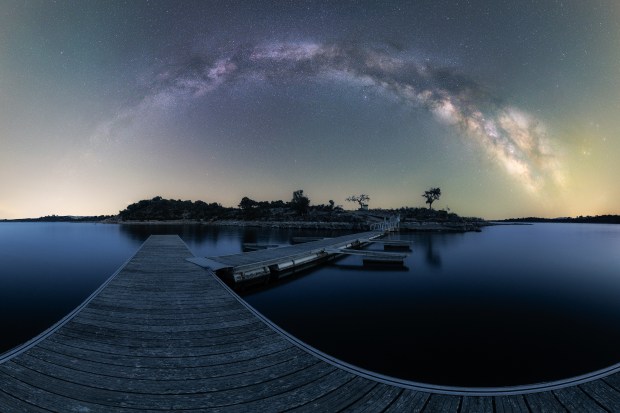 The Milky Way arcs in the sky over a dark blue waterscape with a pier and an island cutting through the center of the scene. Airglow is also visible at both ends of the panorama, a hazy pale green close to the horizon.