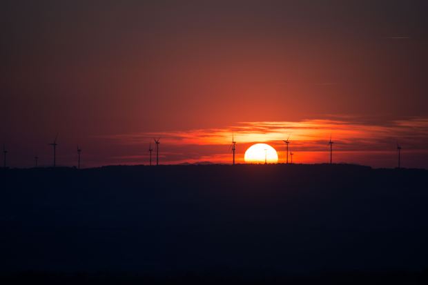 The Sun sets behind a wind farm, casting an orange glow above the horizon and silhouetting wind turbines