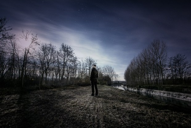 A man stands on a road flanked by winter trees, looking up at a night sky with clouds and stars.