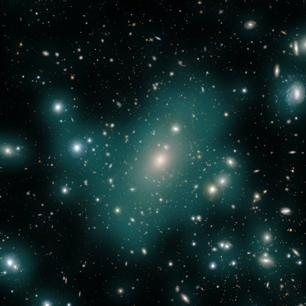 An image of hundreds of galaxies of different sizes and shapes against a black background. A haze of teal gas-like light appears and connects some of the galaxies in the image. The teal glow is intracluster light and is focused mostly towards the middle of the image.