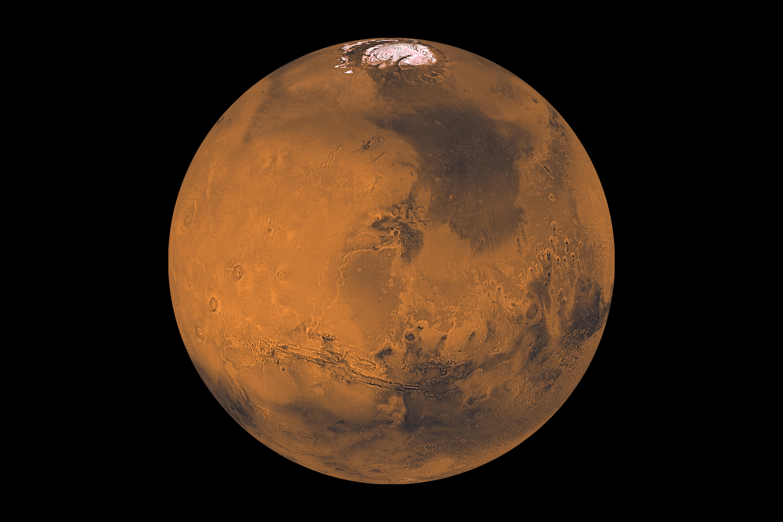 The sandy red orb of Mars is shown against a black background. Numerous dark regions are scattered across its surface, and the bright, white northern polar ice cap is visible at the top of the image.