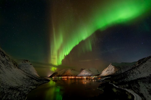 Green ribbons of aurorae arc across the upper two-third of the image, looking like a single wing of an angel (or perhaps the flow of the lifestream from a certain Japanese RPG). Below is a fjord, with sea flanked by snow-covered mountain slopes. The auroral light and light from small villages reflects off the snow.